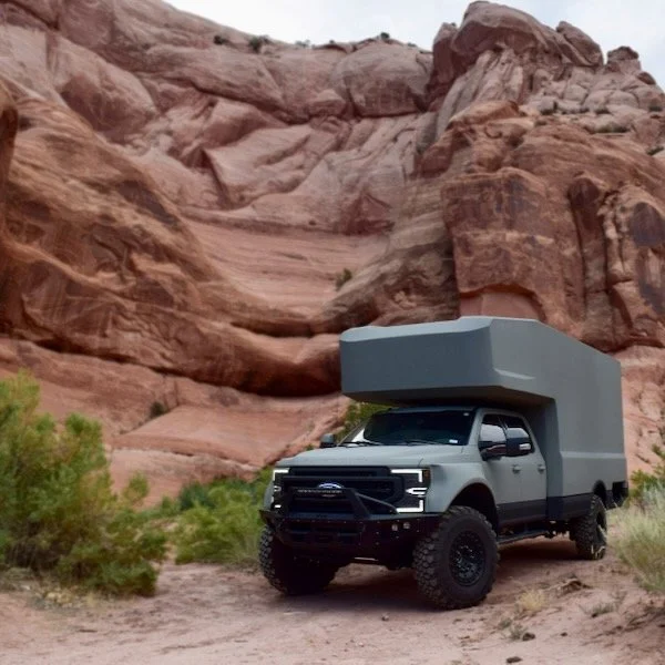 A modified off-road truck with a large, gray rooftop camper parked on a dirt trail in a desert landscape with red rock formations and sparse green vegetation.