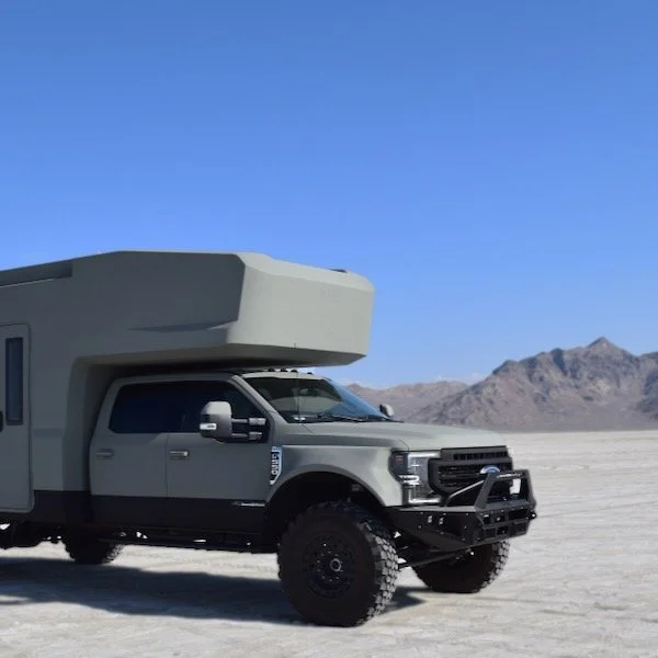 A large gray off-road truck with a camper attachment driving across a flat, desert landscape with mountains in the background under a clear blue sky.
