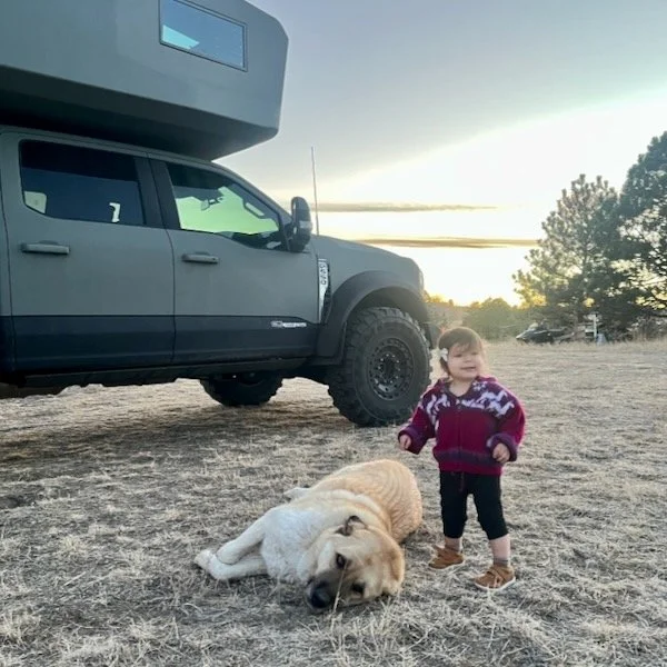 A young child standing next to a reclining golden retriever on a dirt field with a large black truck and camper in the background, during sunset.