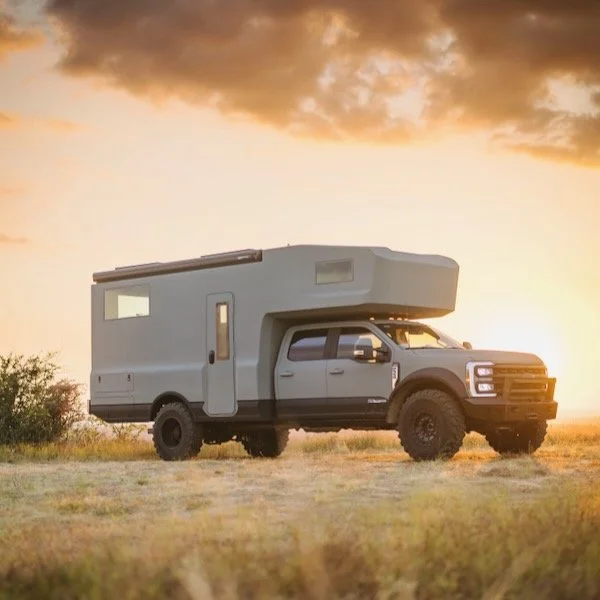 A large truck converted into a camper parked in an open field during sunset with cloudy skies.