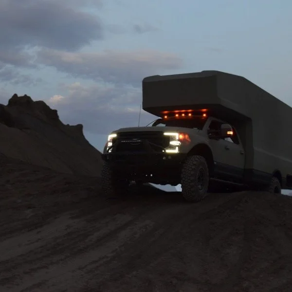 A black pickup truck equipped with a camper shell is parked on a hilltop during dusk or dawn. The truck's headlights are on, illuminating the dark landscape around it, with rocky terrain and sky with clouds in the background.
