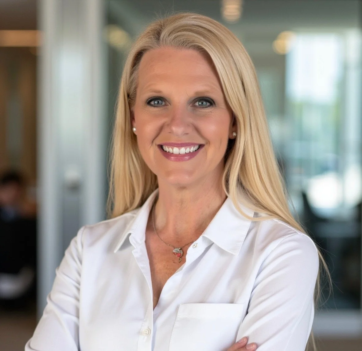 A smiling blonde woman with blue eyes wearing a white blouse and jewelry, standing in a modern office setting.