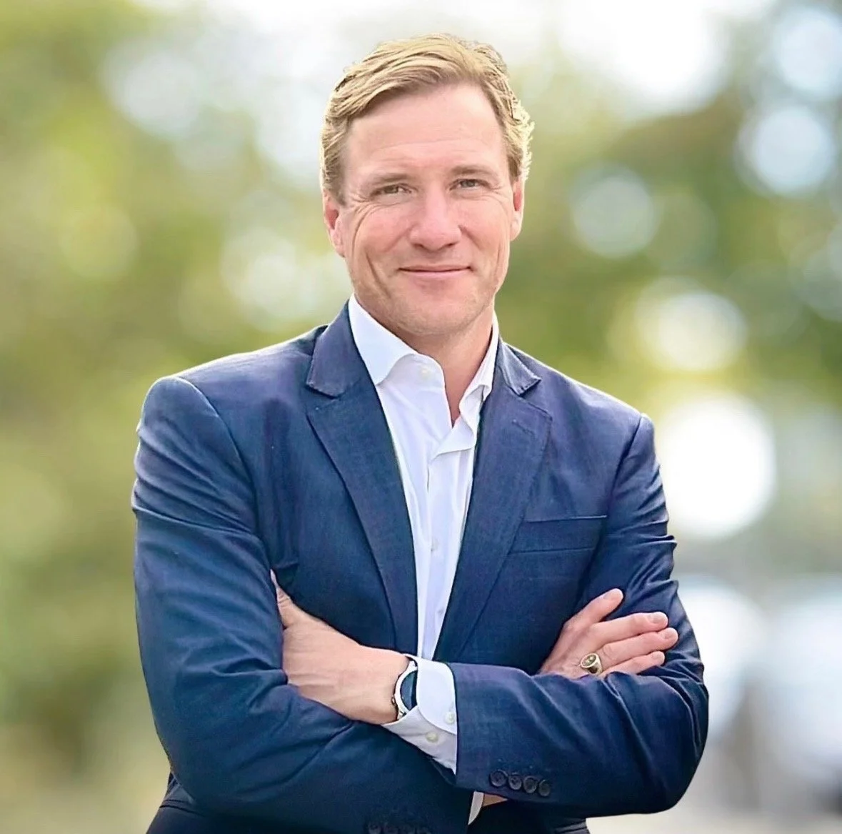 A man in a blue suit with white shirt, standing outdoors with arms crossed, smiling, with blurred green background.