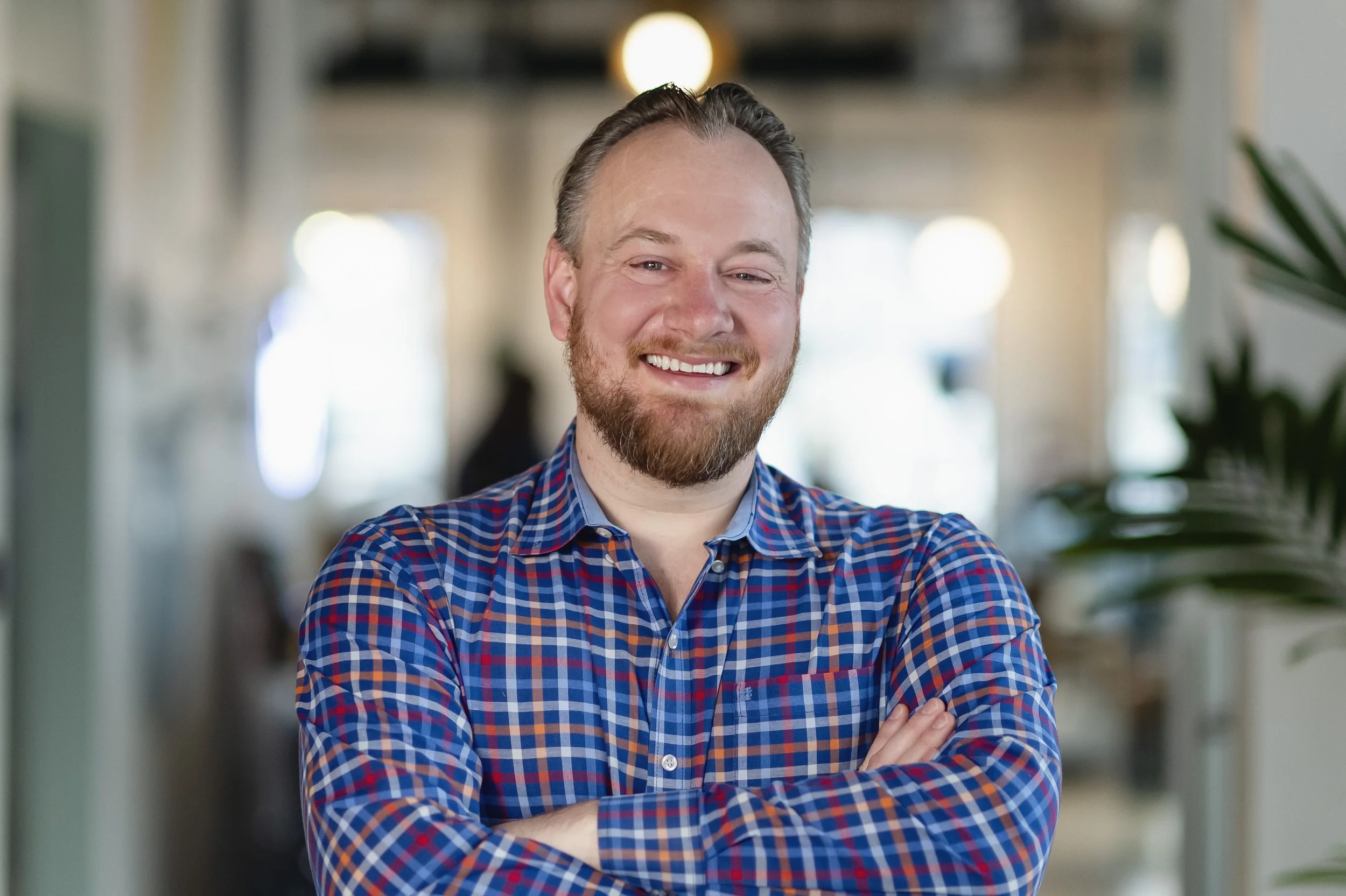 A smiling man with a beard and short hair, wearing a blue and red checkered shirt, standing with his arms crossed in an indoor setting with blurred background lighting and indoor plants.