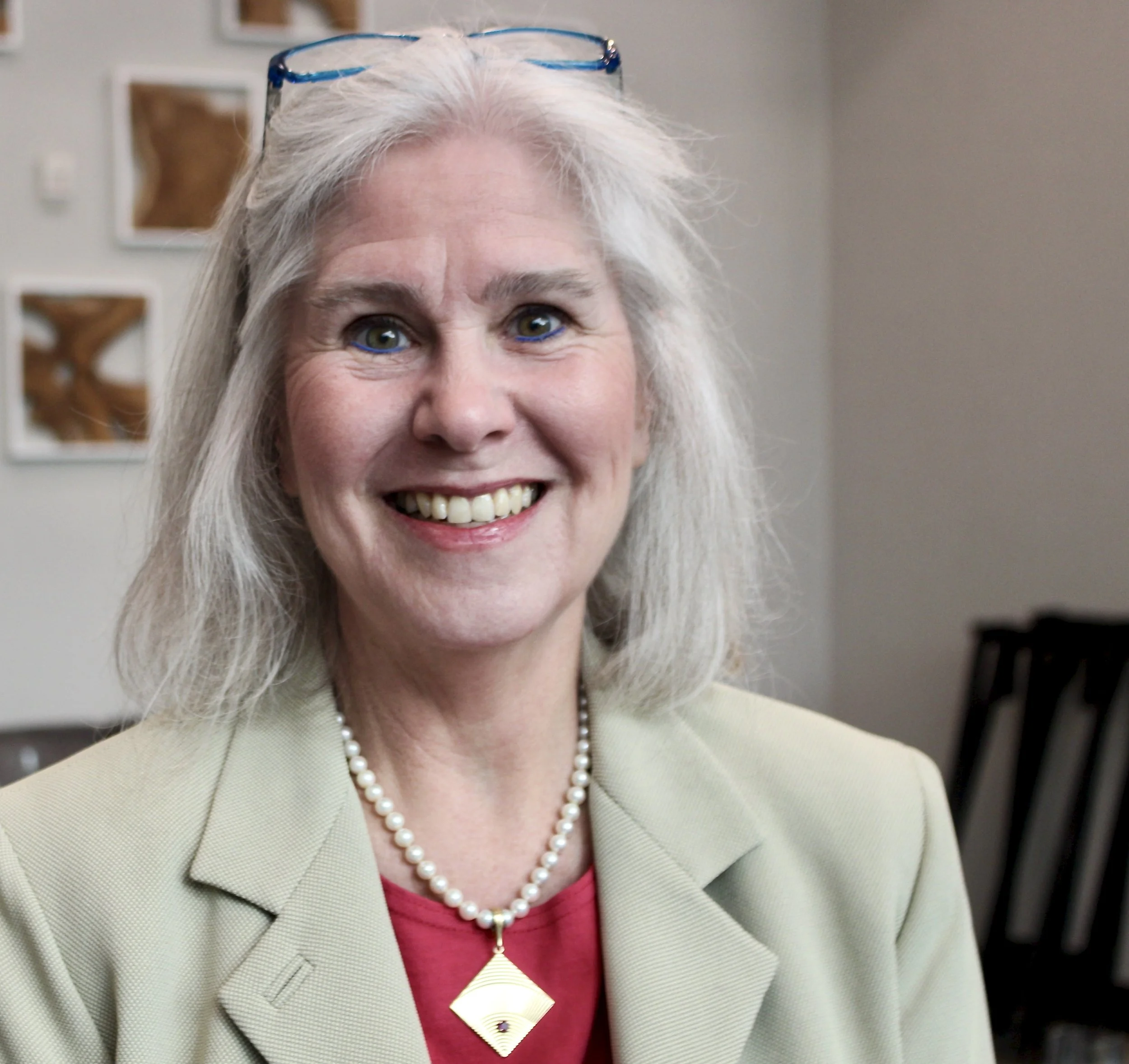 A smiling middle-aged woman with long gray hair, wearing glasses on her head, a pearl necklace with a gold pendant, a red top, and a beige blazer, sitting indoors with art frames on the wall behind her.