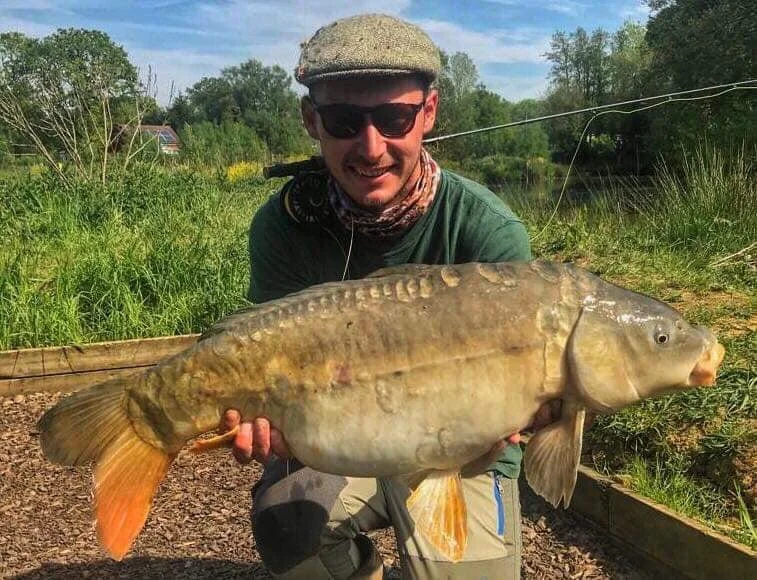 A person wearing a cap and sunglasses holding a large fish, possibly a carp, with a natural outdoor background.