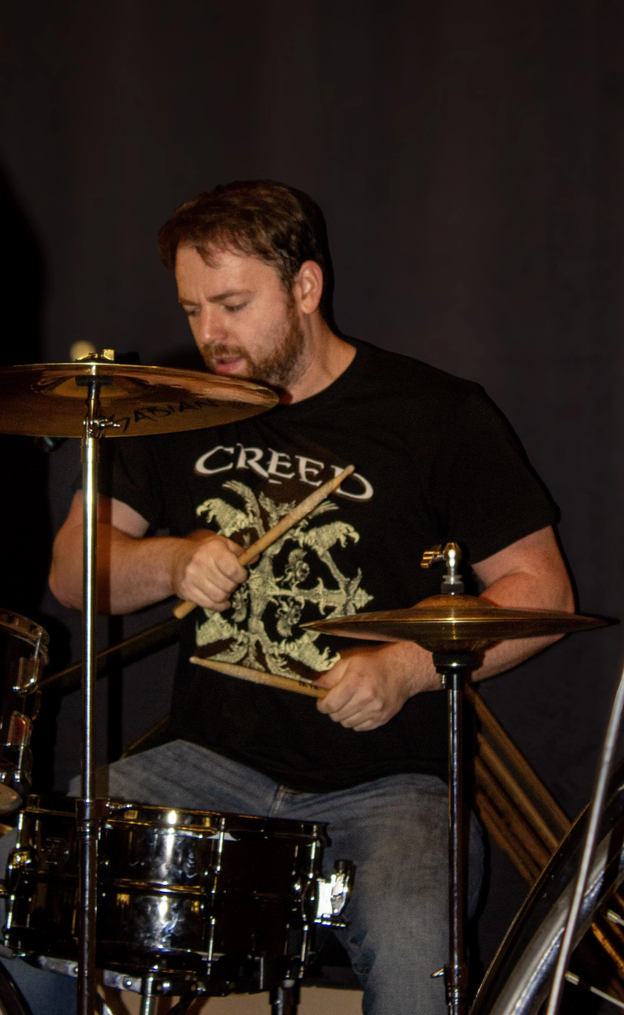 A man playing drums, wearing a black T-shirt with the word 'CREEP' and a graphic design, with a dark background.