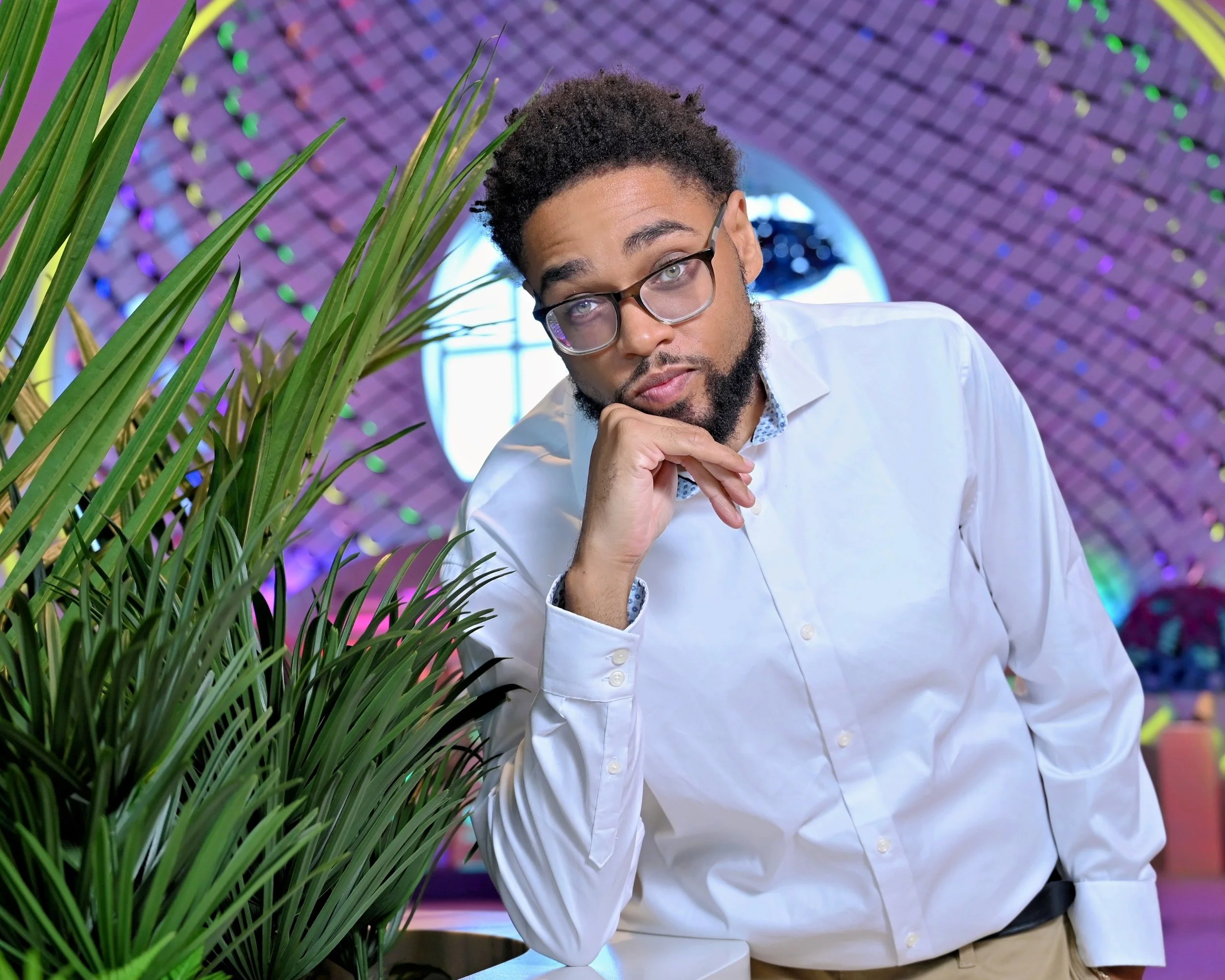 A man with glasses, a beard, and curly hair posing with one hand on his chin in an indoor space with colorful, illuminated decor, and green plants.