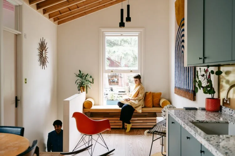 A woman sitting on a window seat in a modern, light-colored living space with wooden ceiling beams, a potted plant, a decorative wall hanging, a red rocking chair, and a kitchen area with blue cabinets.