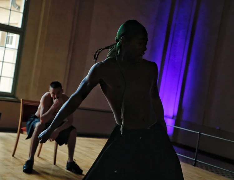 A shirtless man with a bandana and a woman with a dark ponytail performing in a dance or theatrical scene on a stage with purple lighting and wooden floors.