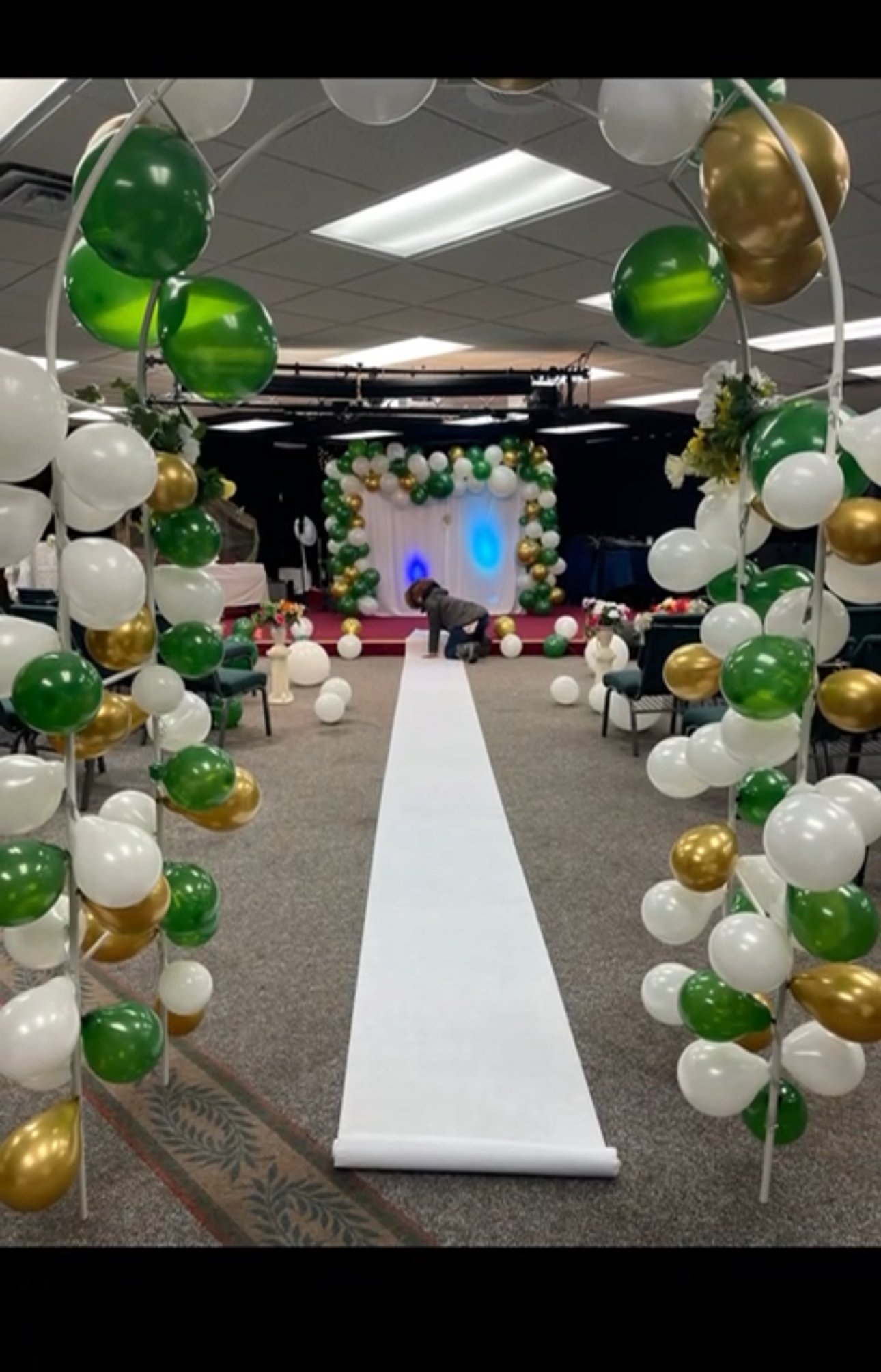 Event venue setup with white, green, and gold balloons and a central white aisle leading to a decorated stage.