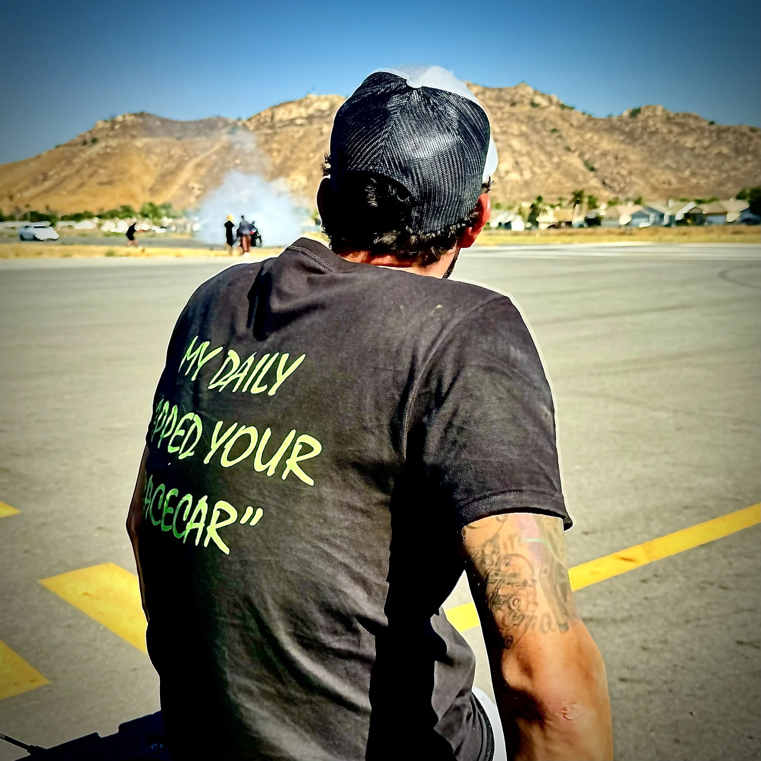 A man wearing a black cap and dark t-shirt sitting on a motorcycle, watching a burnout during a car event on a clear day with mountains in the background.