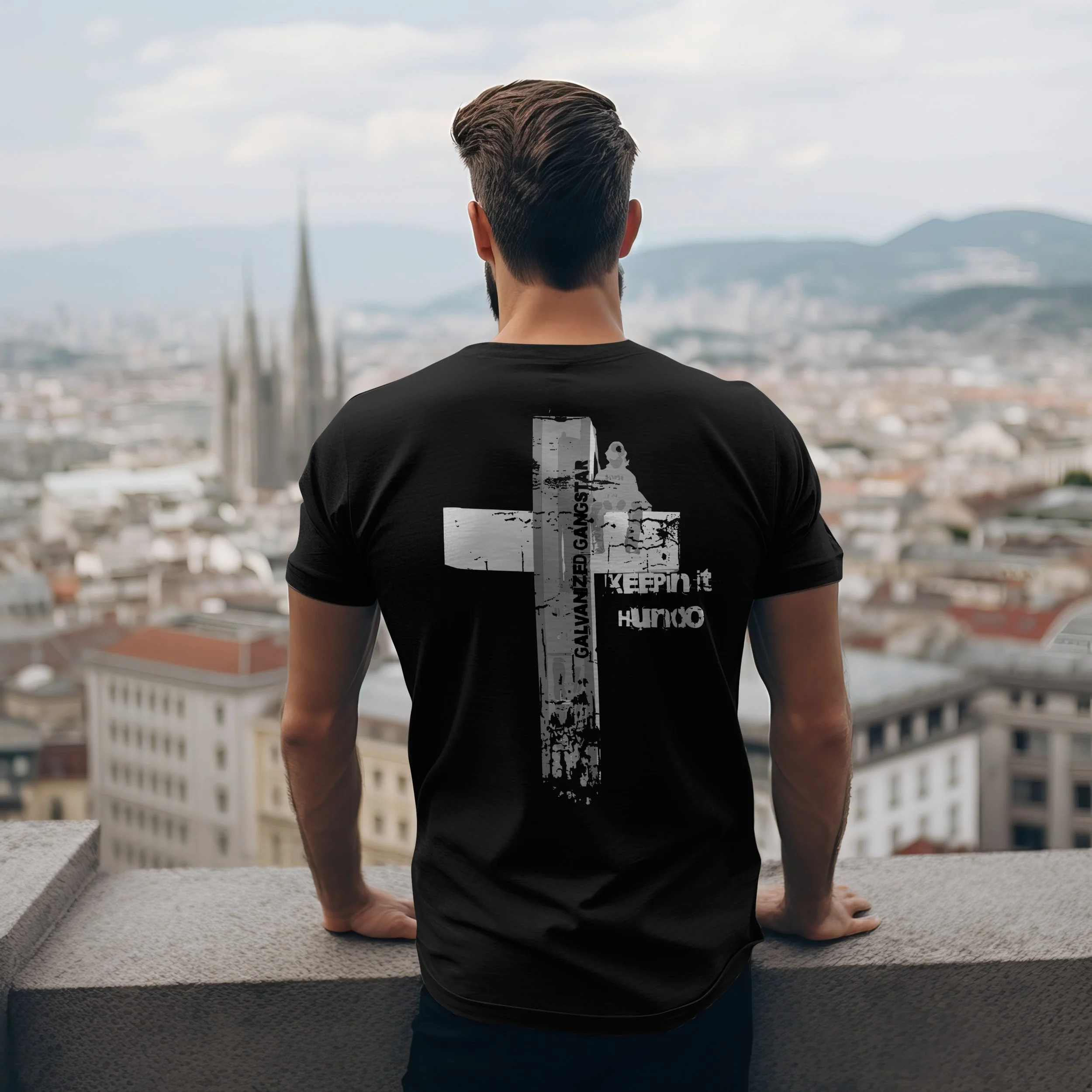 Man with styled hair wearing a black T-shirt with white graphic text and bold cross design, standing on a rooftop overlooking a cityscape with a cathedral and mountains in the background.