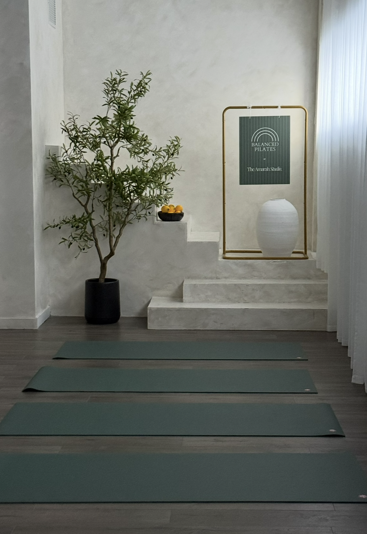 A yoga studio with three yoga mats on the wooden floor, a potted plant on the left, a bowl of lemons on stairs, a standing sign with 'Balanced Pilates' and 'The Amarah Studio,' a large white vase, and a white curtain on the right.