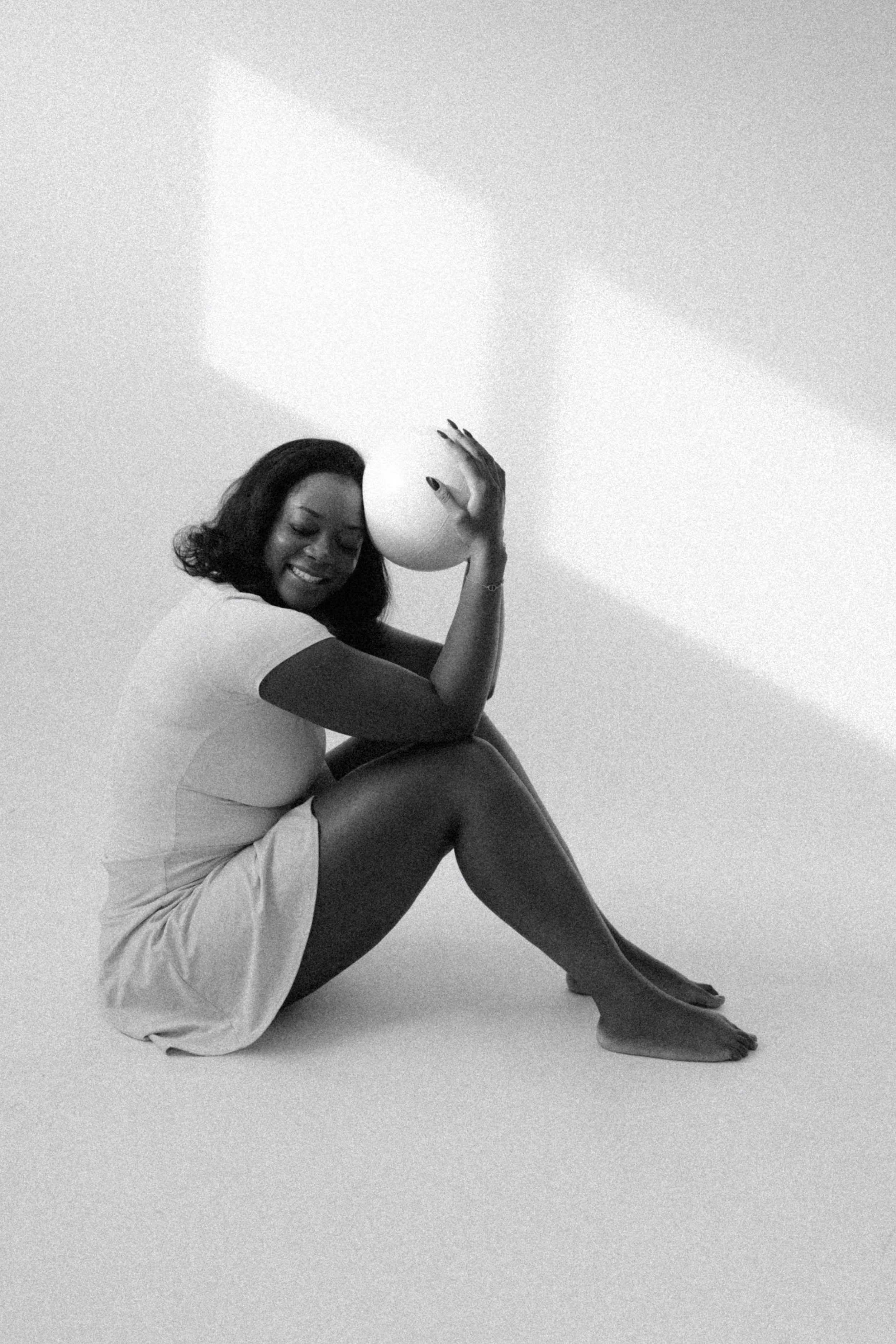 A black and white photo of a woman smiling, sitting with one knee raised and holding a balloon near her head.