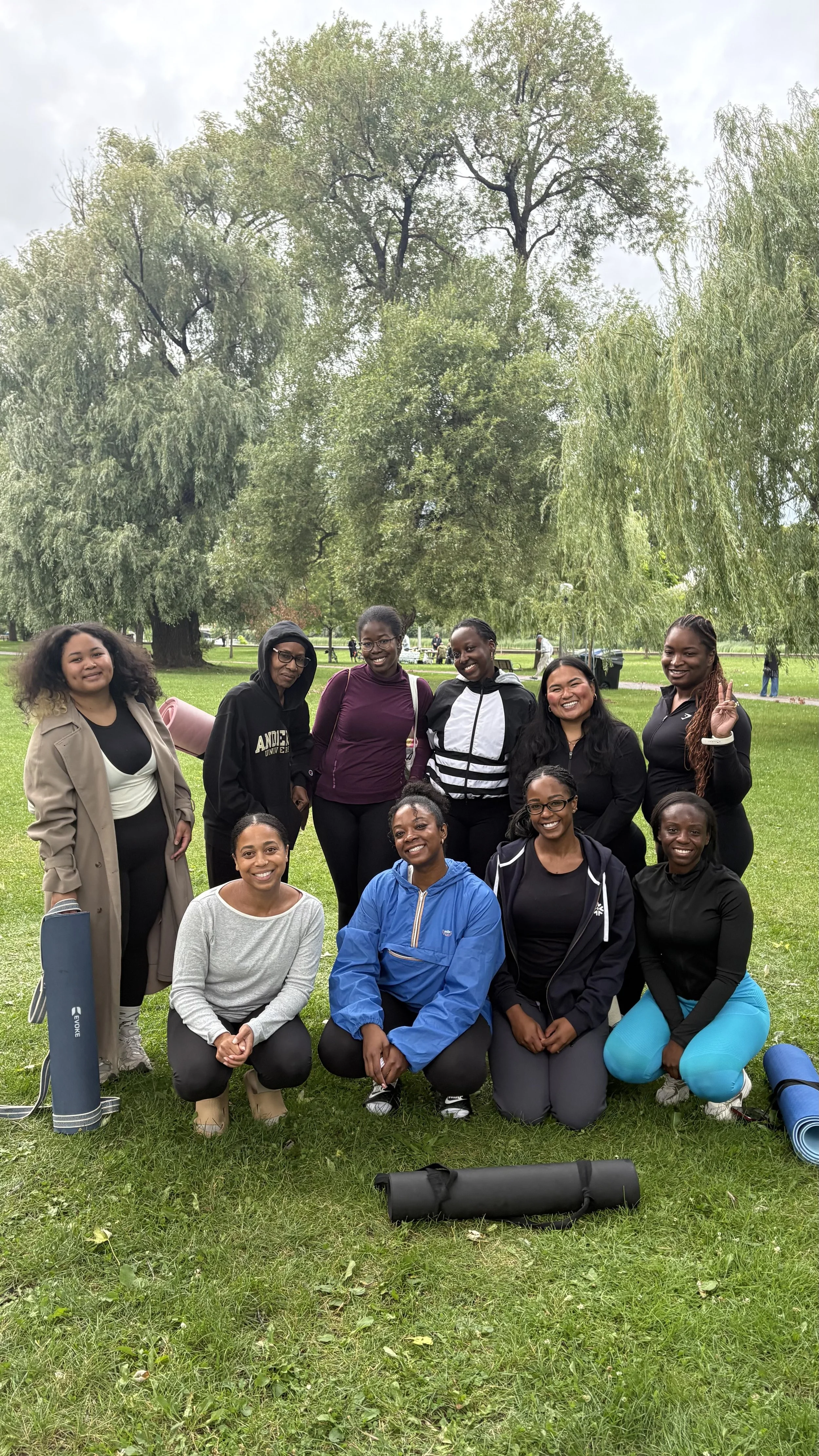A group of ten women of diverse ethnicities posing together outdoors in a park with green grass and large trees in the background. Some are standing, others are kneeling or sitting. They are dressed in casual athletic wear, and there are yoga mats and a foam roller on the ground in front of them.