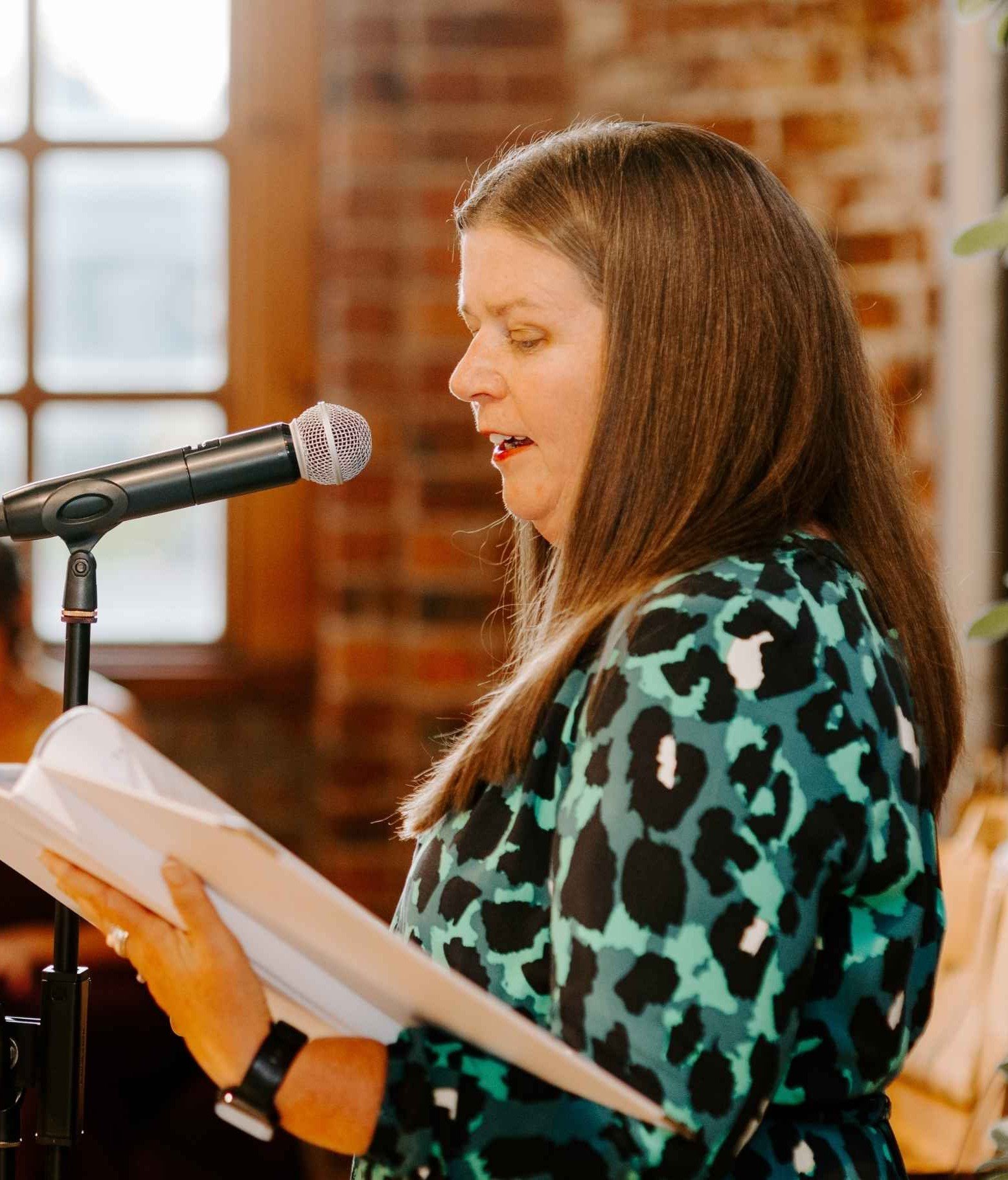 Kate is wearing a patterned dress at an autumn wedding. She is both reading the story of how the couple came to be wed, and convening an award ceremony for the friends and family who supported them on the way.
