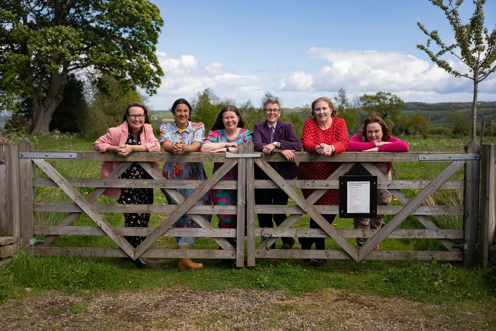 Six women are leaving over a wooden gate to a natural burial site on a bright spring. They are smiling at the camera.