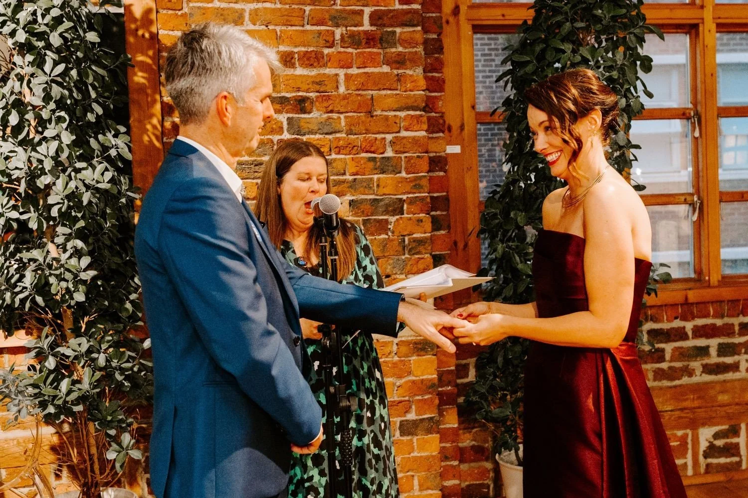 The bride, wearing a long burgundy dress, smiles at the blue suited groom while placing a wedding ring on his finger. Kate, in a blue and green animal print dress reads the unique promises written by the bride.