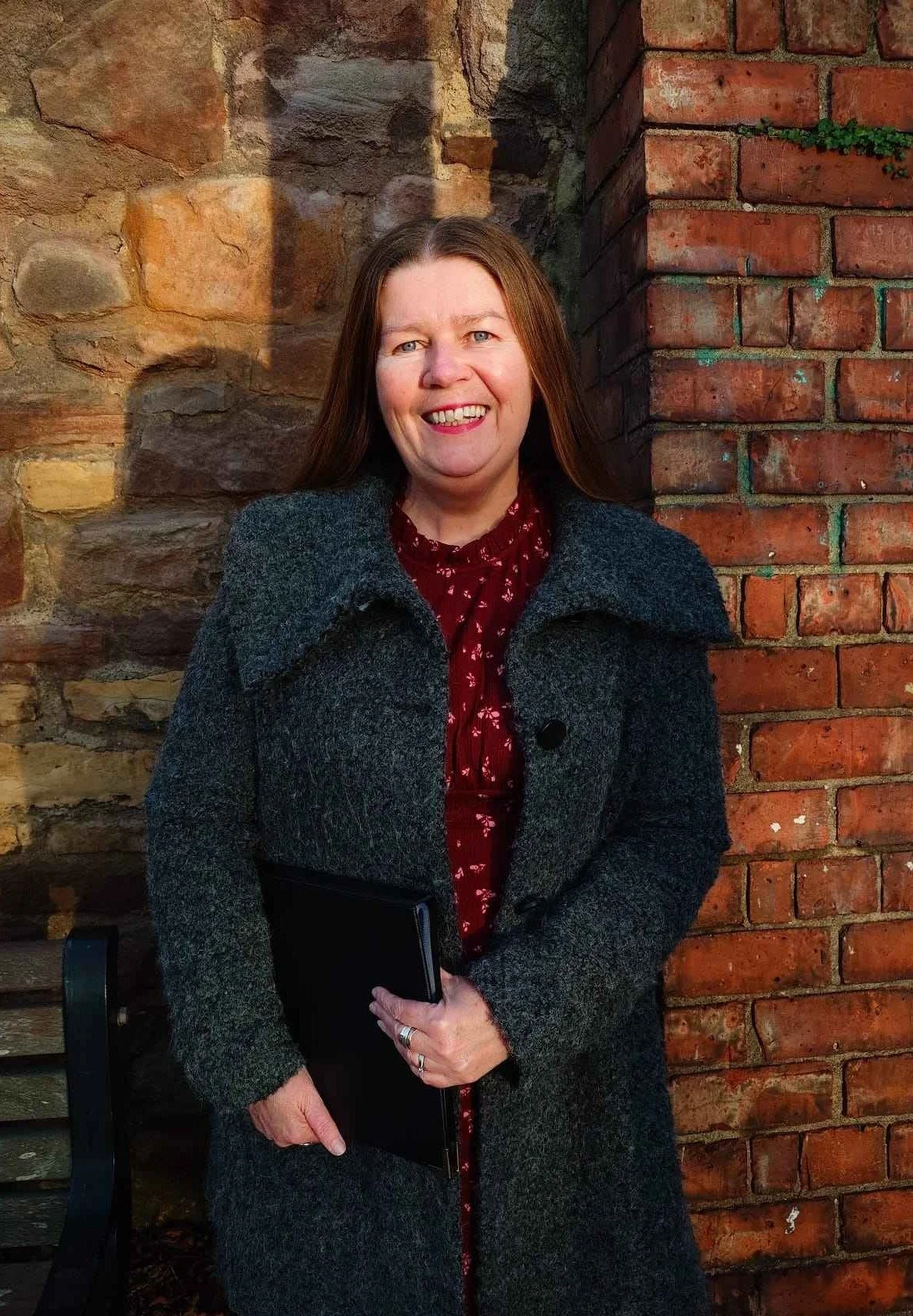 Kate, a memorial ceremony celebrant in Newcastle is a woman with long brown hair. She smiling and holding a black ceremony folder outside a community venue used for a memorial service in Newcastle. The venue has beautiful brick and stone walls.