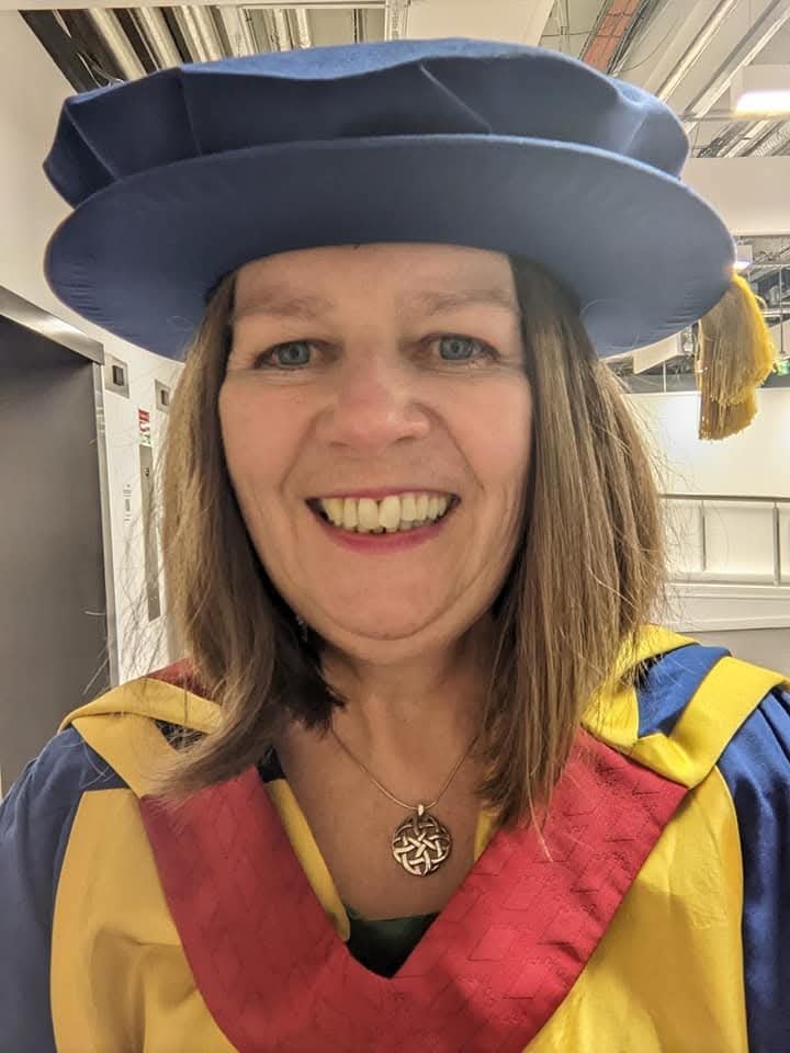 A smiling Kate is wearing her doctoral robe which is a royal blue academic cap and a royal blue, yellow and red gown.