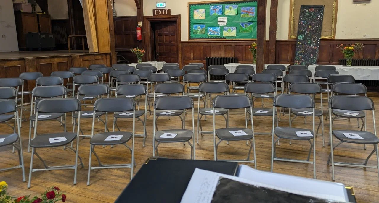 This image was taken at a memorial service in a community centre. There are wooden panels on the walls and wooden floors. There are vases of flowers around the hall. Rows of seats in one half of the space  are visible although there were 120 seats.