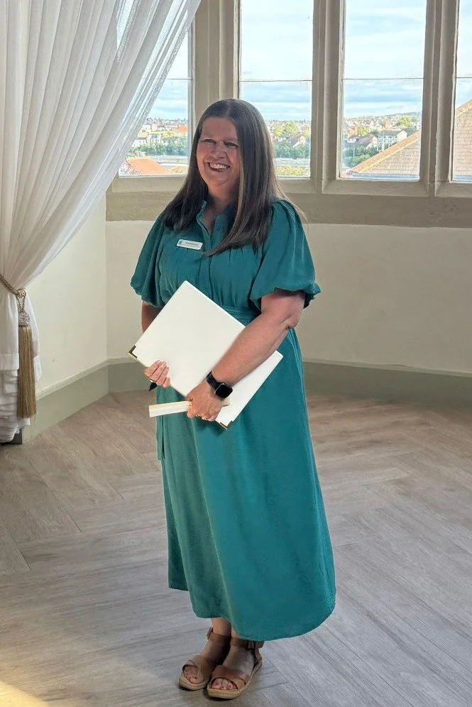 This photo of Kate was snapped in the seconds before the start of a wedding as the bride had arrived. She is wearing a long teal dress and holding a white script folder and a white fan. She is smiling widely.