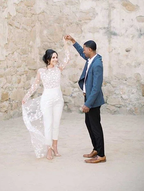 This image, available from Sourced and Co shows a newly wed couple holding hands aloft. The man is wearing a blue suit jacket and dark trousers. The woman is wearing a white outfit will a lace cape.