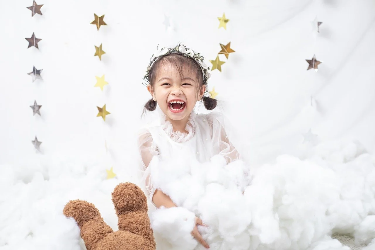 A laughing child wearing a white dress has a teddy in front of her. There are gold star decorations in the background.