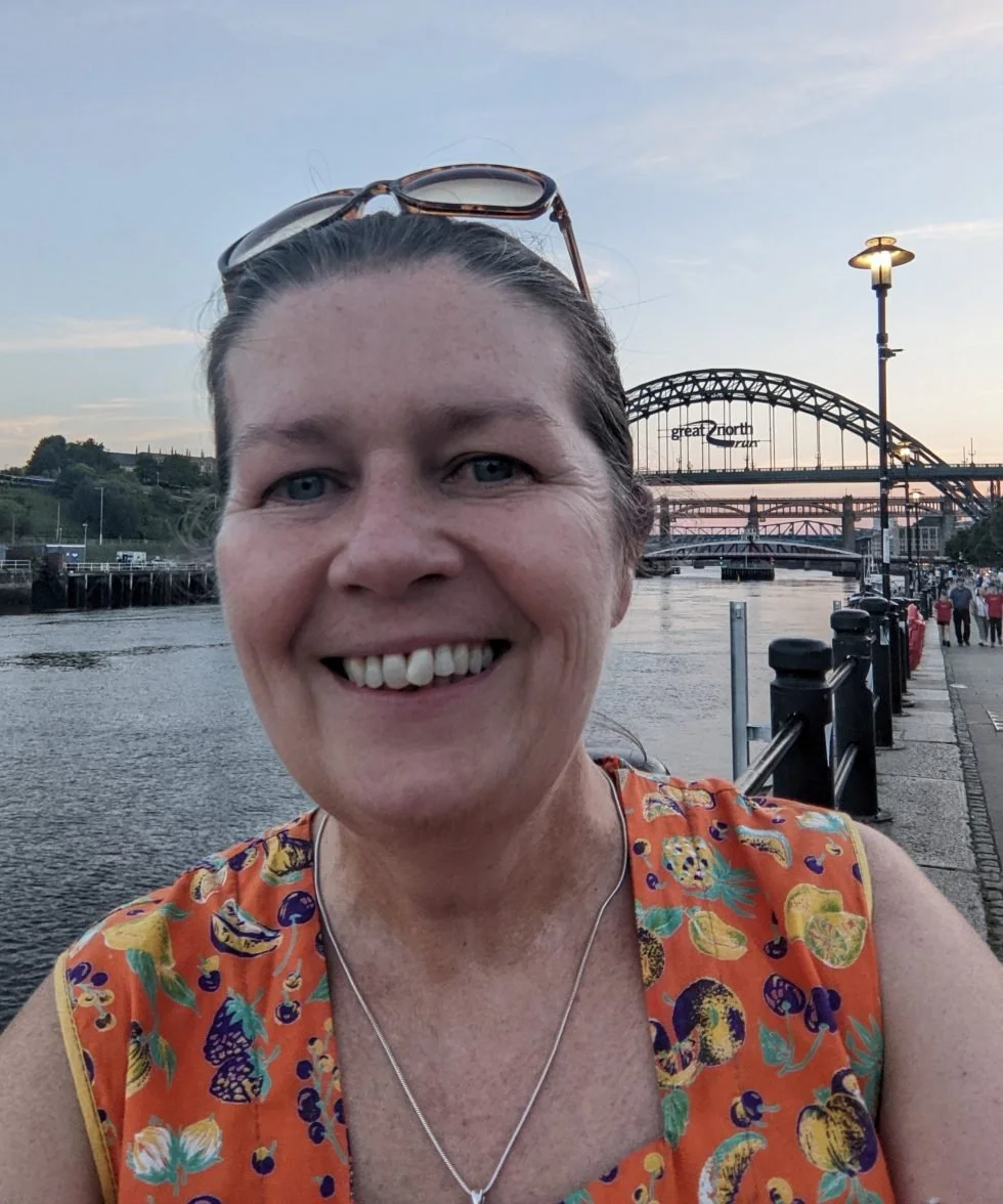 Kate is standing beside the River Tyne in Newcastle on a sunny evening. She is wearing an orange dress and smiling. The Tyne Bridge is visible in the background with the Great North Run sign. The Swing and High Level Bridges are also visible.