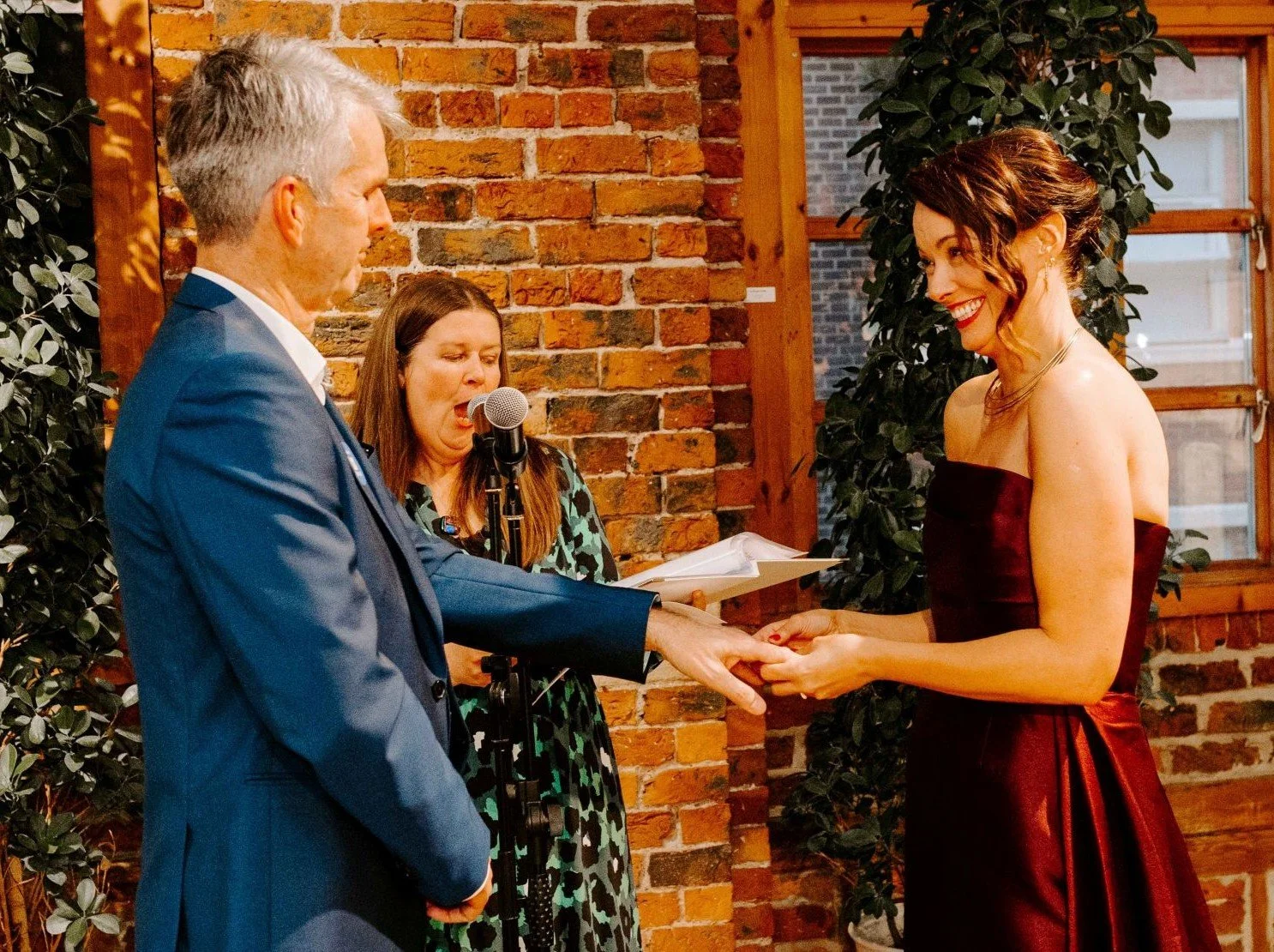 The bride, wearing a long burgundy dress, smiles at the blue suited groom while placing a wedding ring on his finger. Kate, in a blue and green animal print dress reads the unique promises written by the bride.