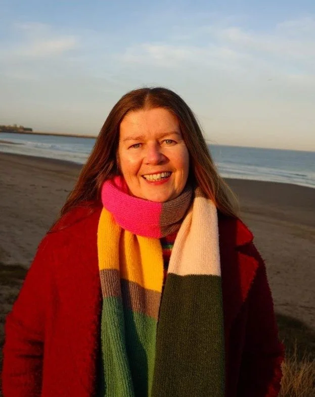 Image of Kate wearing a berry coloured winter coat, multi-coloured scarf and striped rainbow jumper standing at South Shields Beach in the sunshine of a winter afternoon