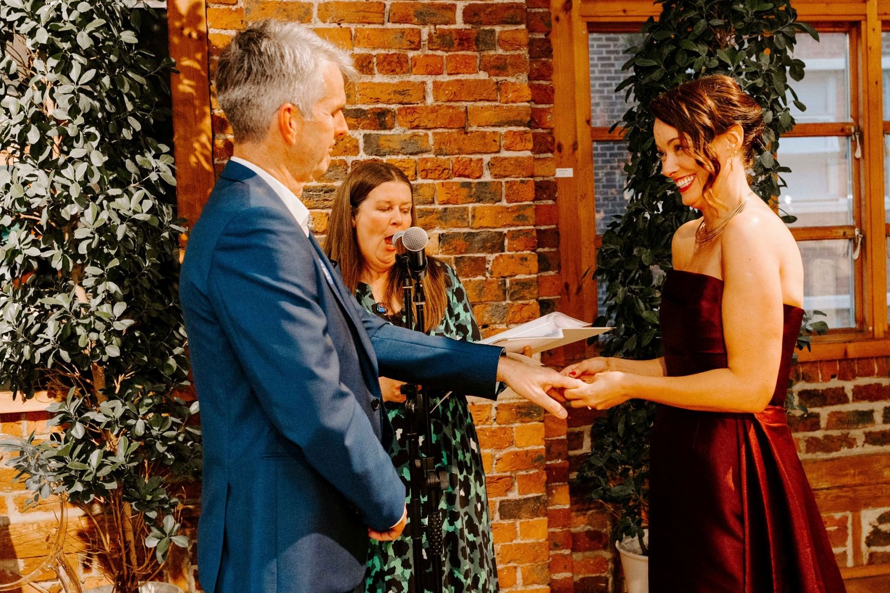 The bride, wearing a long burgundy dress, smiles at the blue suited groom while placing a wedding ring on his finger. Kate, in a blue and green animal print dress reads the unique promises written by the bride.