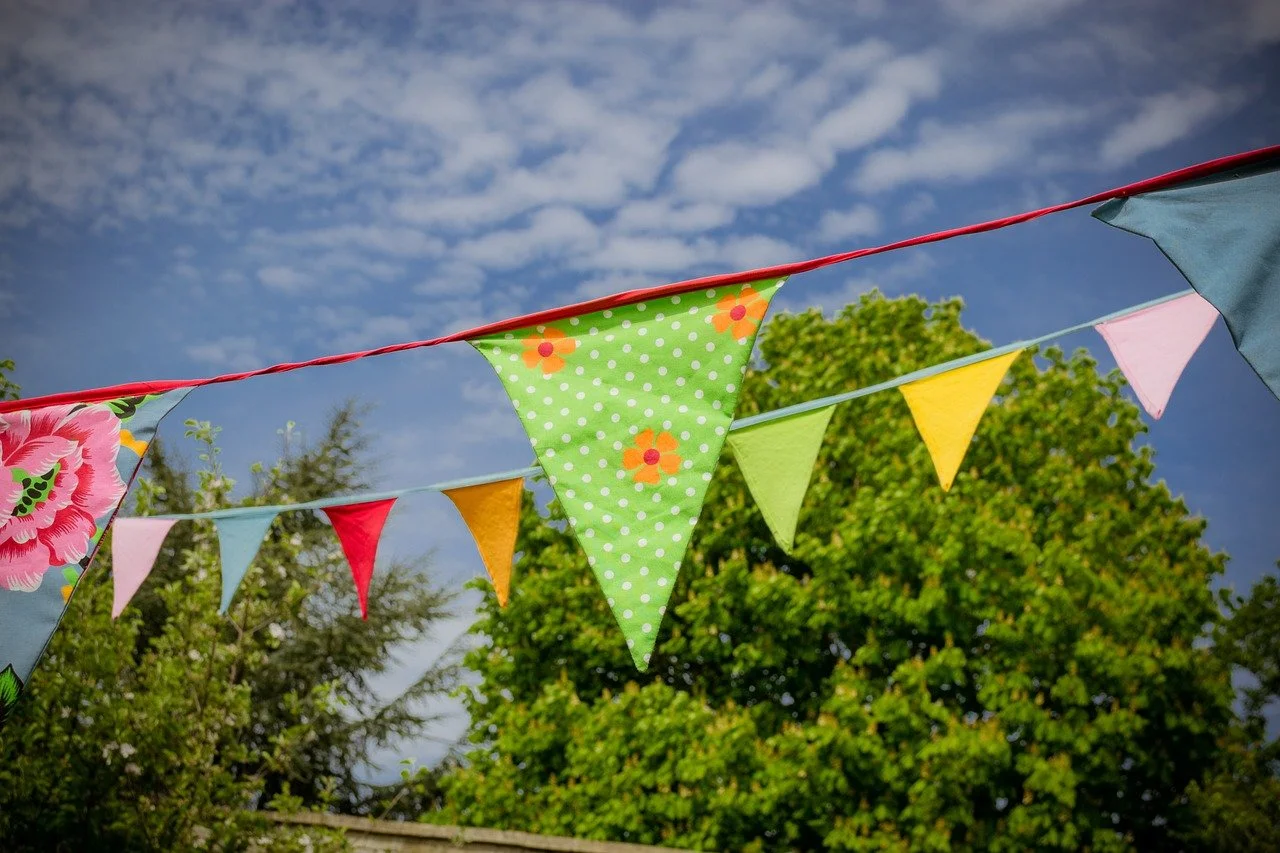 Colourful fabric bunting hanging outdoors against green trees and a blue sky.