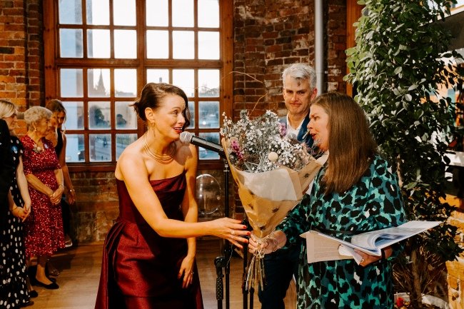 A bride in a burgandy dress presents a bouquet of flowers to Kate in a blue leopard print dress. The groom in a blue suit looks on smiling.