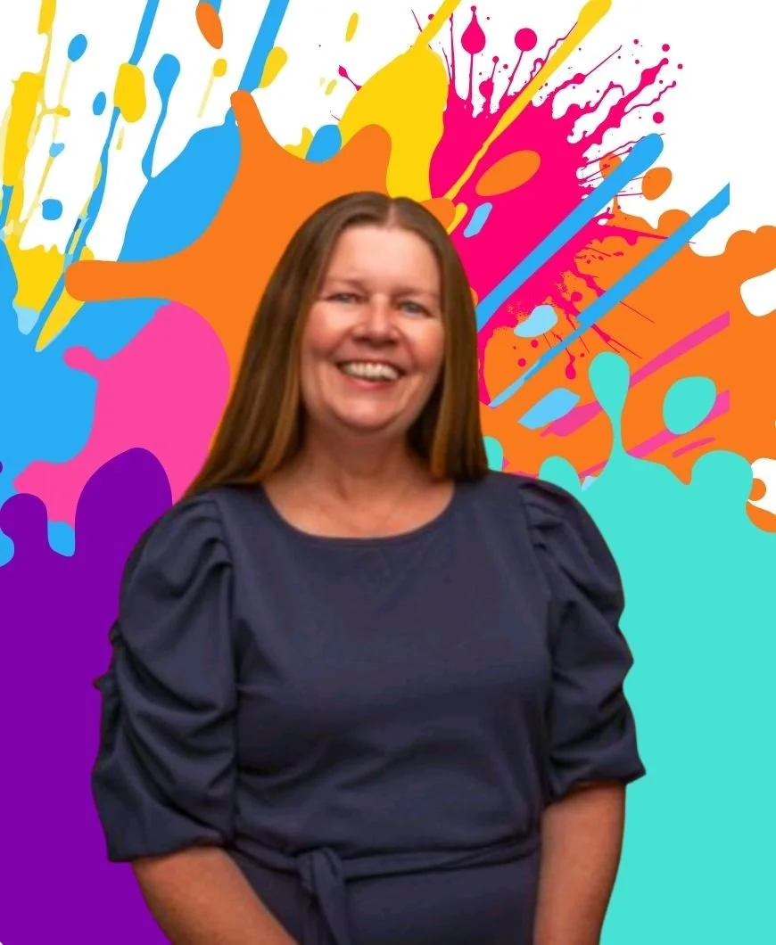 In this image Kate is wearing a dark blue dress and is smiling in front of a colourful paint splash effect background. There are purple, orange, yellow, pink and jade blue colours in the background.