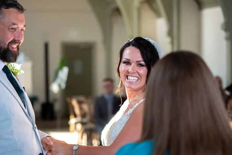 In this image the back of Kate's head is visible. The nearly weds, a woman with black hair and a white dress and a man with dark beard and a cream suit are holding hands, looking at Kate and smiling.