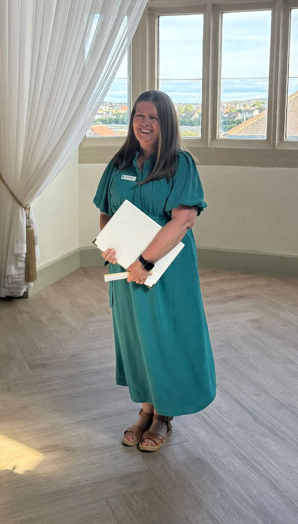This photo of Kate was snapped in the seconds before the start of a wedding as the bride had arrived. She is wearing a long teal dress and holding a white script folder and a white fan. She is smiling widely.