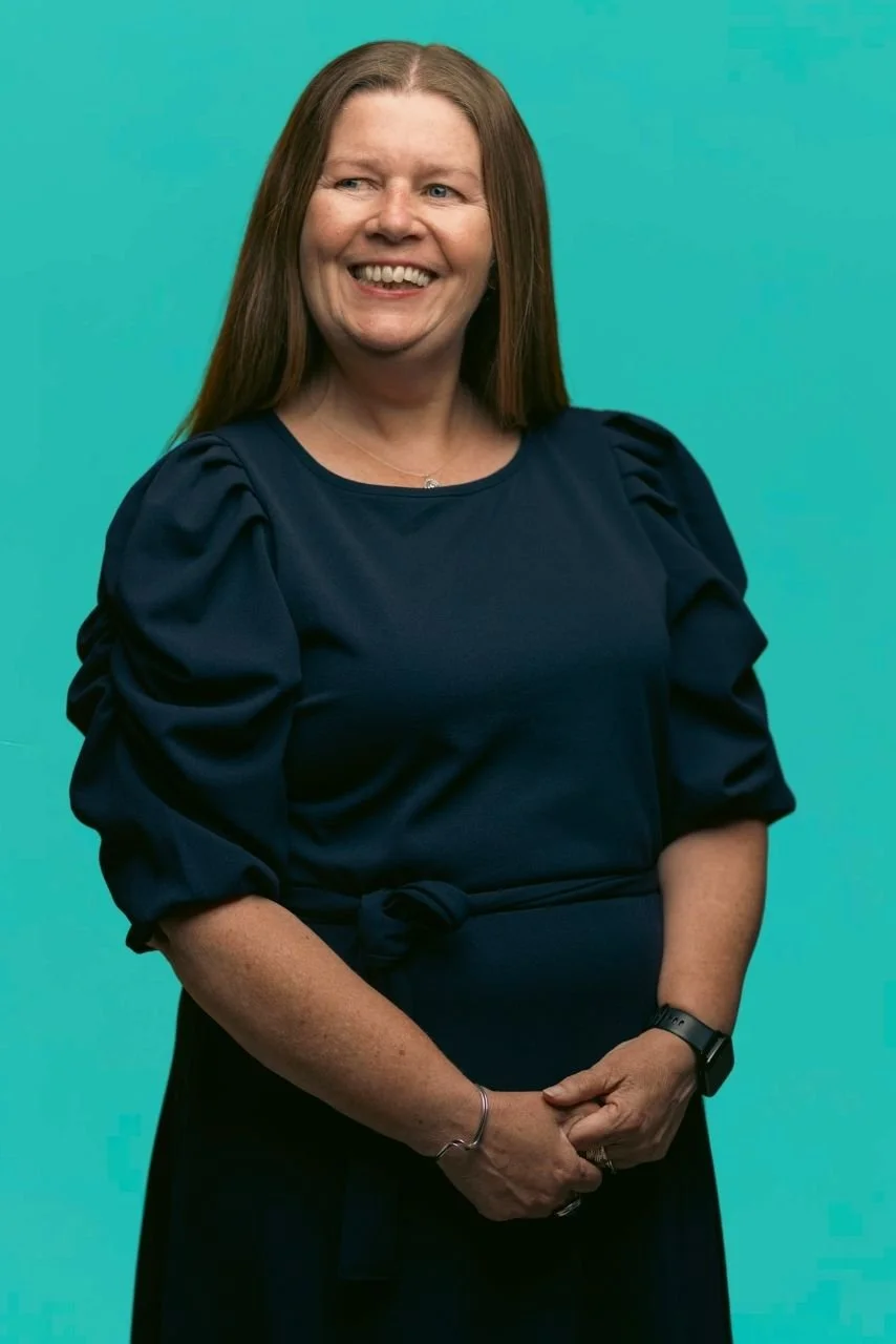 Kate is wearing a blue dress, has her hands clasped in front of her and is smiling widely.