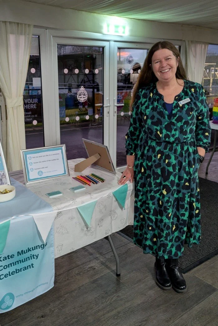 Kate is at a community event at South Shields Football Club. She is standing next to a display table which has bunting, items sitting on a white cloth and a sign that says Kate Mukungu Community Celebrant.