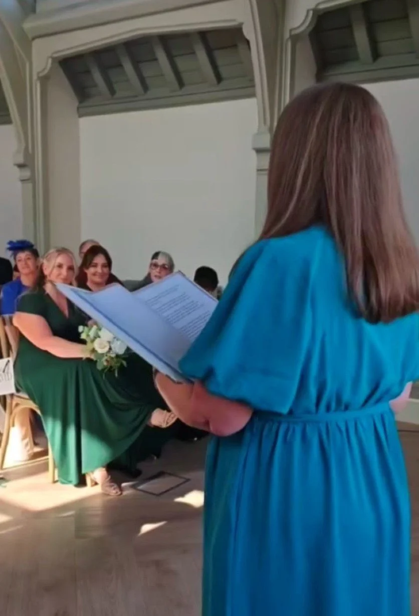 The image of Kate officiating a wedding is taken from a side angle looking out at the guests. Kate is wearing a teal dress, which you can see at the back and is reading from her script.