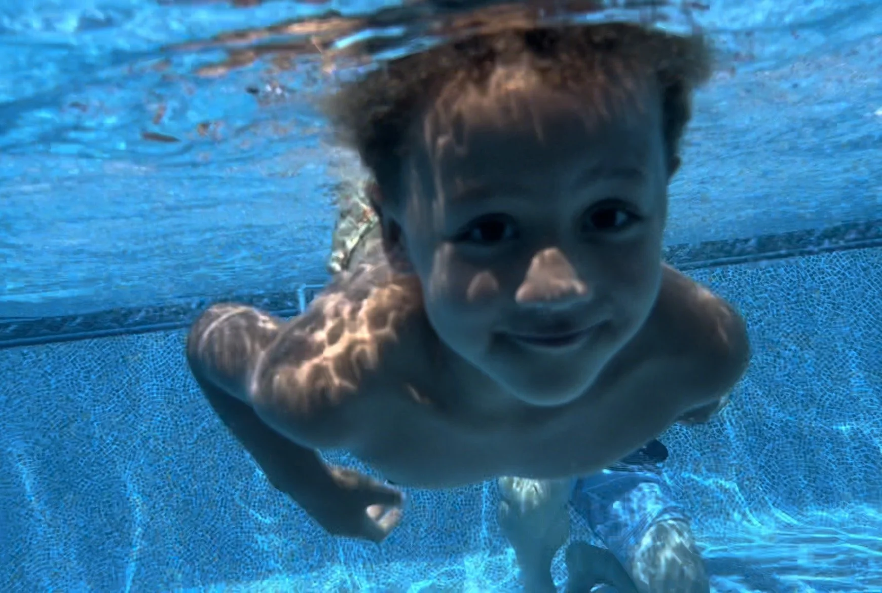 A young boy swimming underwater in a pool, looking at the camera with a smile.