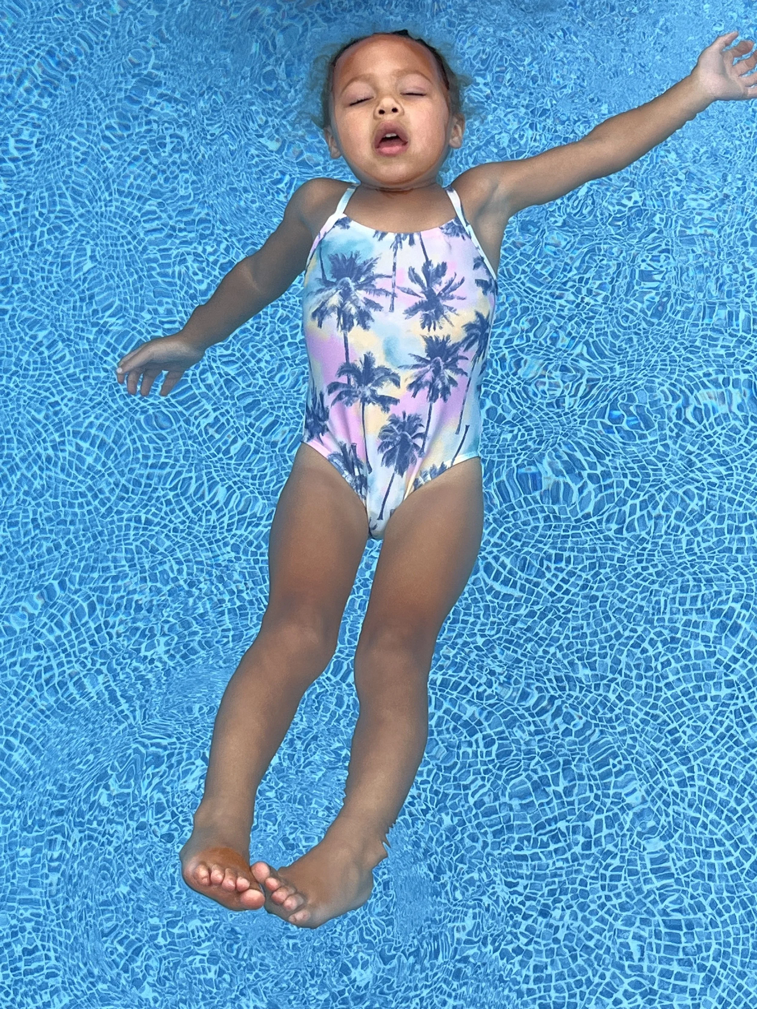 Young girl floating on her back in a swimming pool, wearing a swimsuit with a palm tree print.