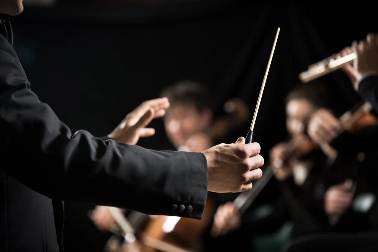 Close-up of an orchestra conductor's hands holding a baton, directing musicians in the background playing instruments.