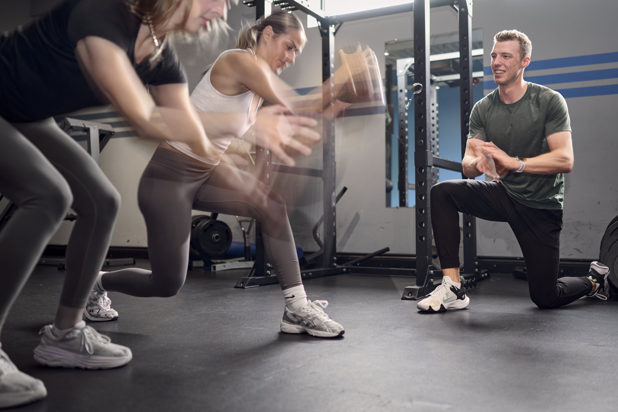 A fitness trainer coaching two women during a workout session at a gym, the two women are performing medicine ball slams.