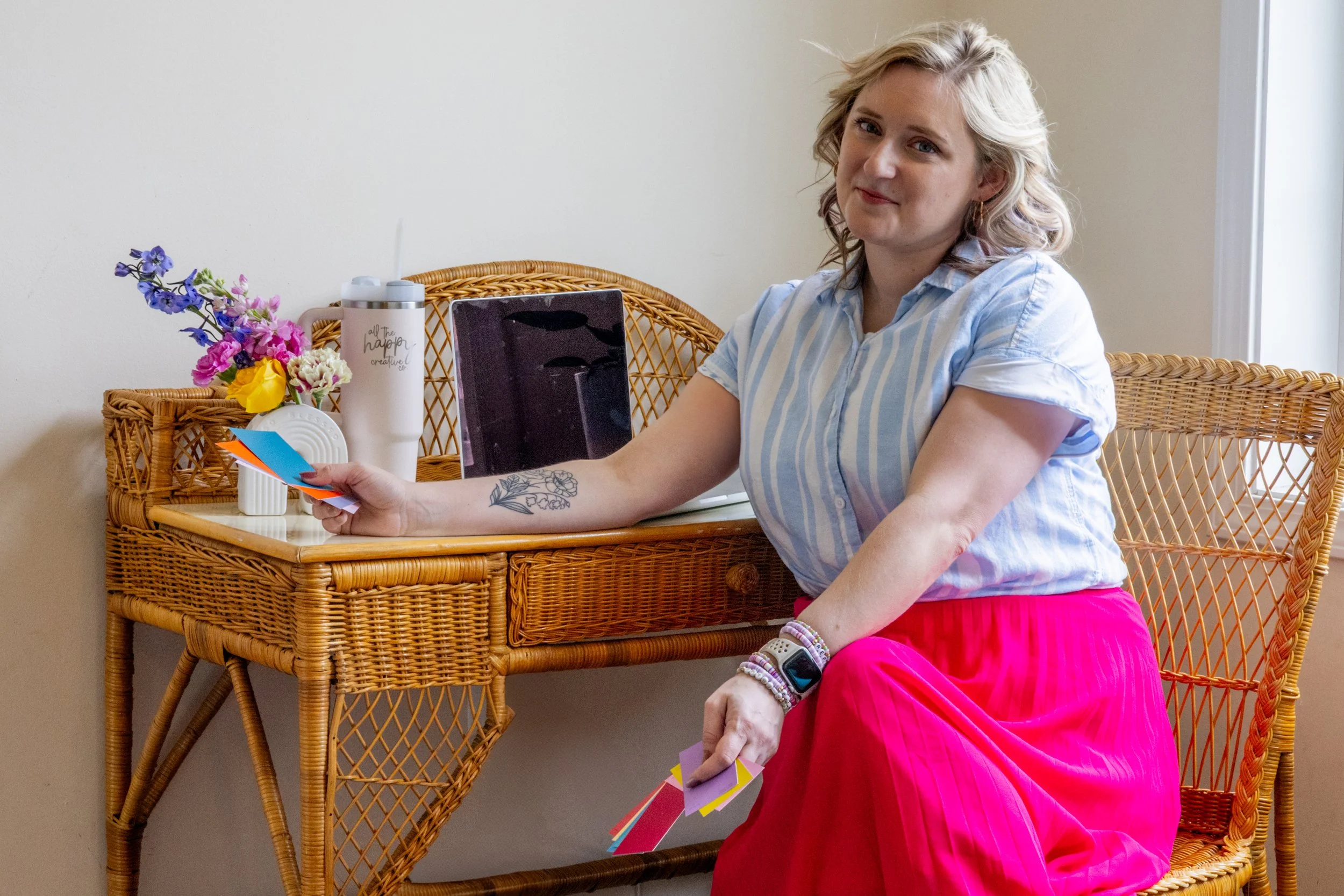 Woman sitting on a wicker chair wearing a striped shirt and pink skirt, holding color swatches. A wicker table beside her holds flowers, a tumbler, and a photo or book.
