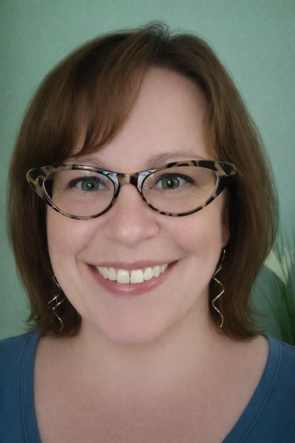Close-up portrait of a woman with short brown hair, wearing patterned glasses and hoop earrings, smiling against a green background.