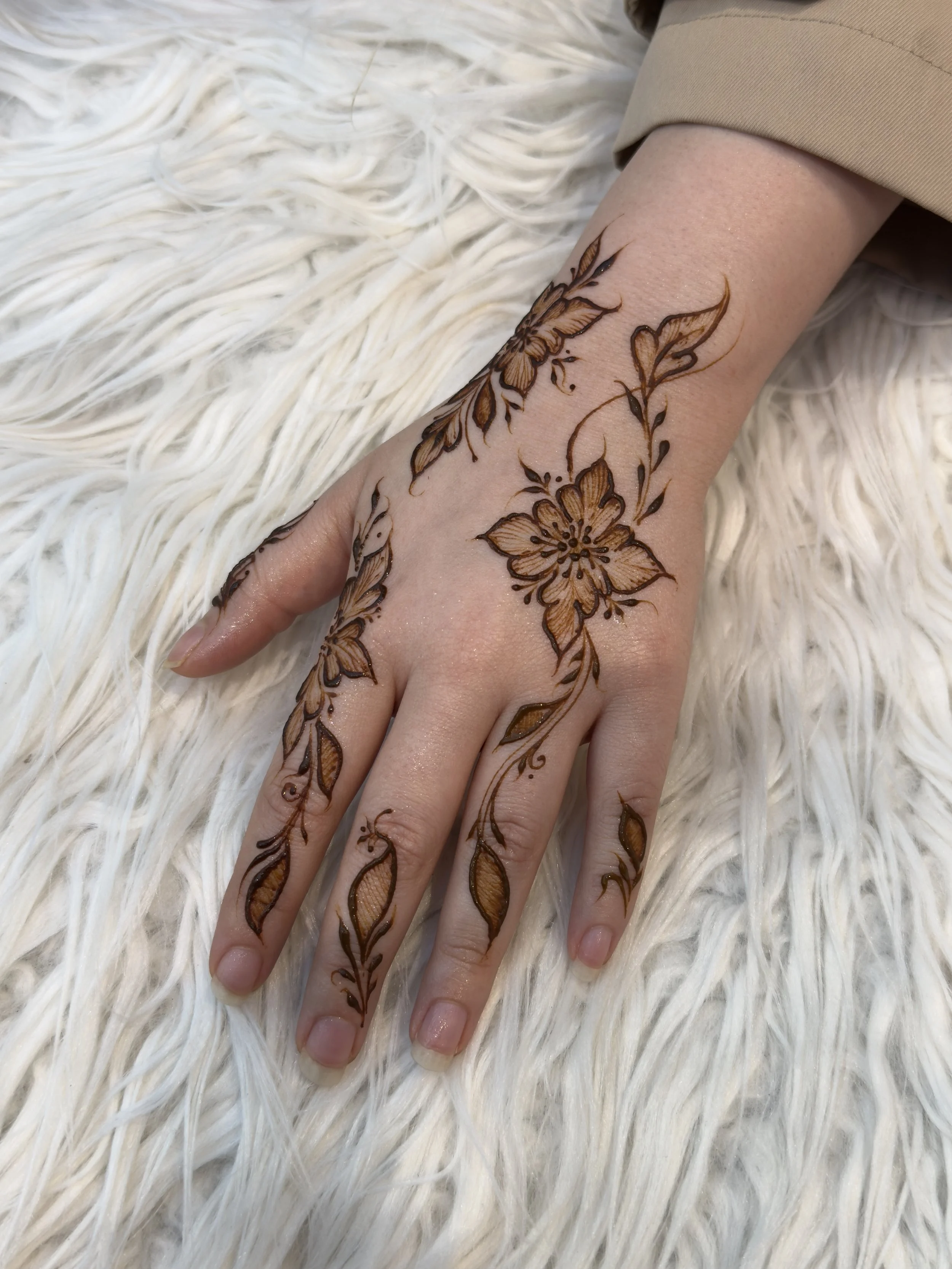 Henna-style floral tattoo on a person's hand and wrist, featuring large flowers with detailed petals and leaves, resting on a white, fluffy surface.