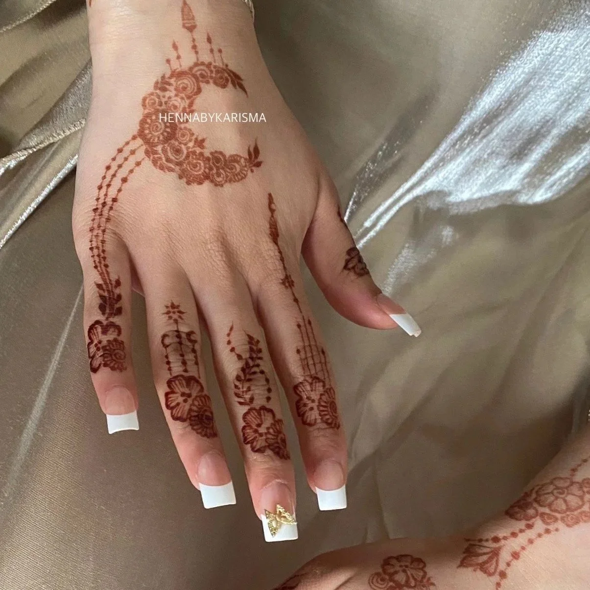 Hand with intricate henna designs, featuring floral patterns and a crescent moon on the back of the hand, against a shiny, metallic fabric background.