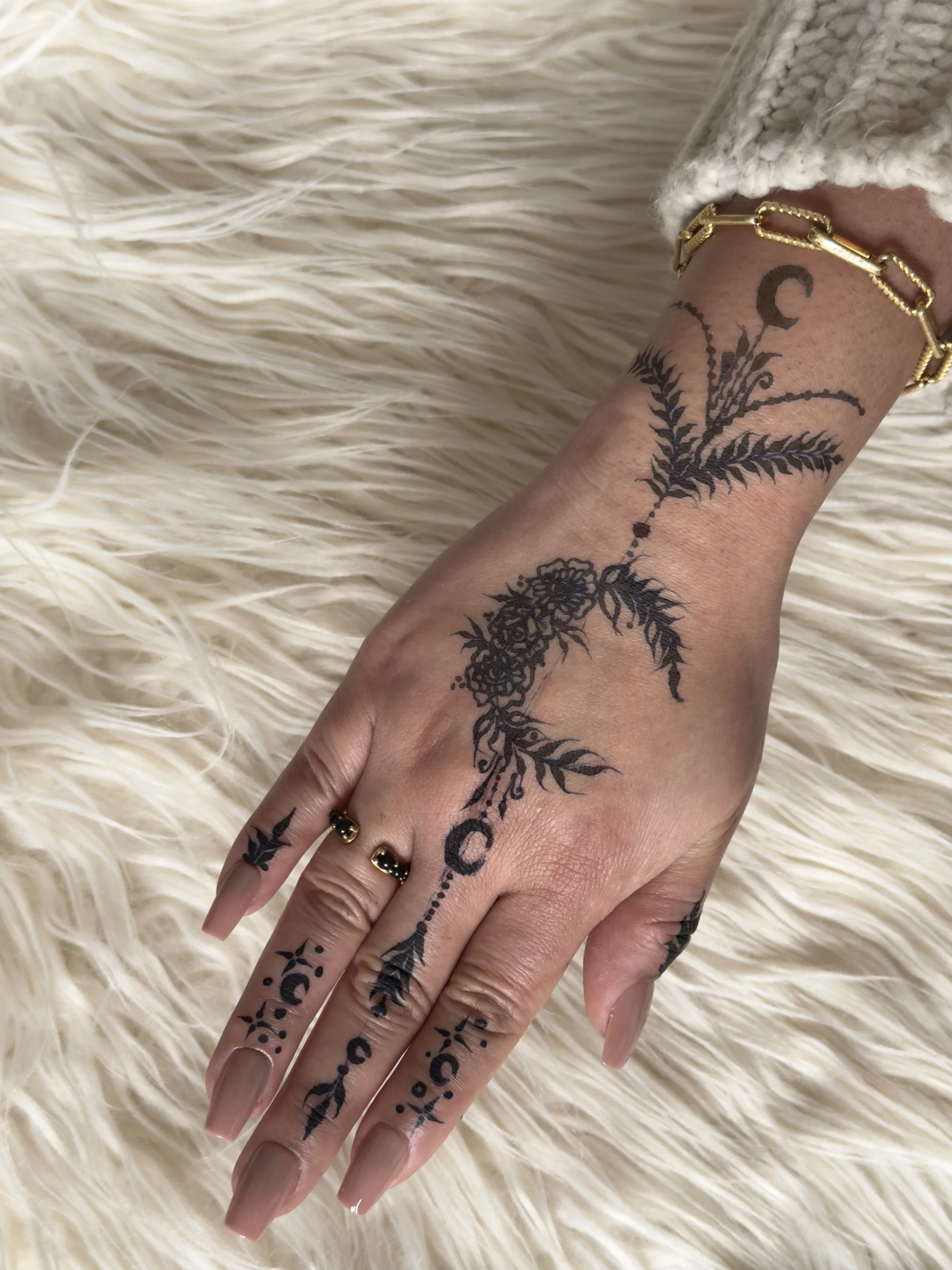 A hand with black tattoos of floral and botanical designs, with gold jewelry including a bracelet and rings, resting on a cream-colored furry surface.